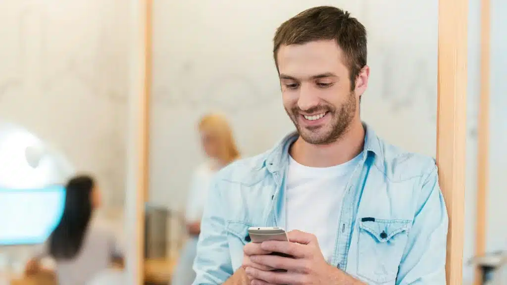 A man smiling while texting on his phone while two women can be seen in the background.