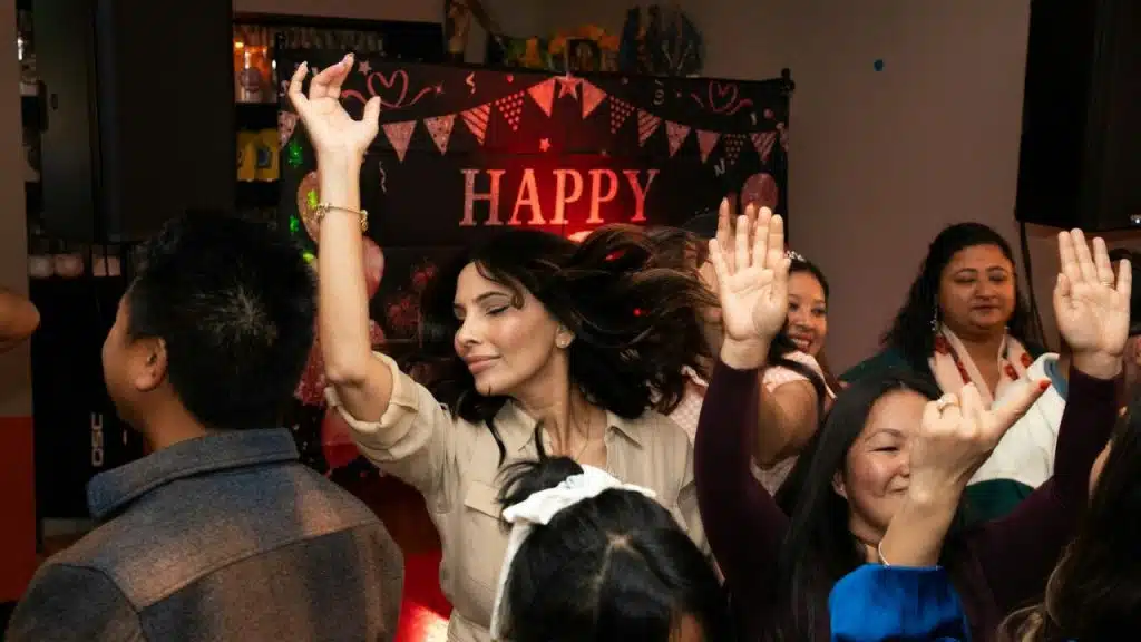 Woman with long dark hair dancing and raising her hands at an indoor party.