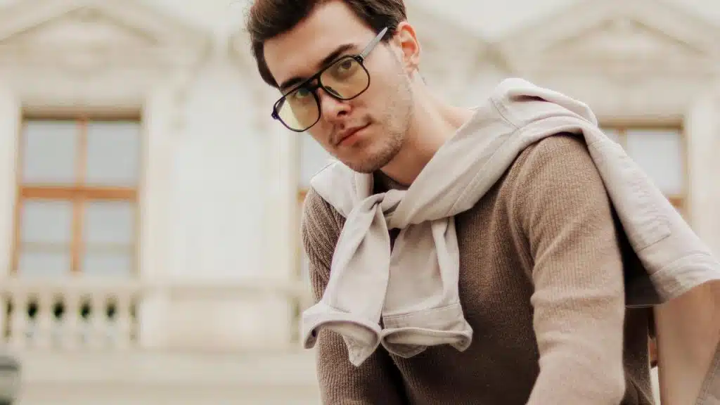 A young man in glasses and a sweater with a jacket tied over his shoulders.