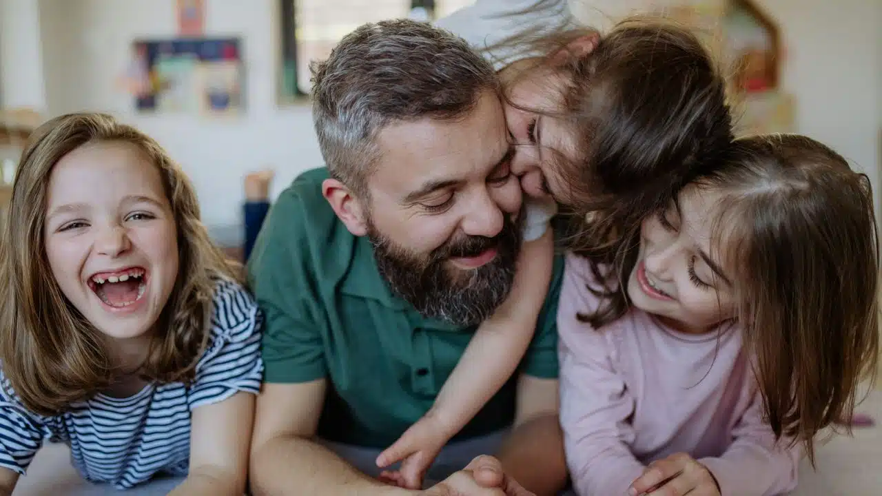 A father with a beard and green shirt is lying down, smiling while being hugged by his three young daughters.