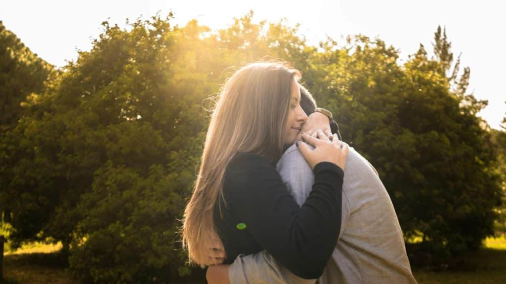 A woman and a man hugging outdoors with sunlight shining through trees.