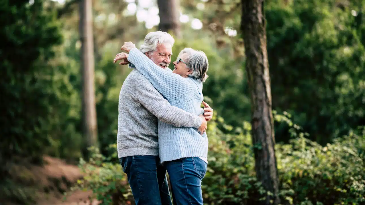 An old couple embrace each other in a forest.