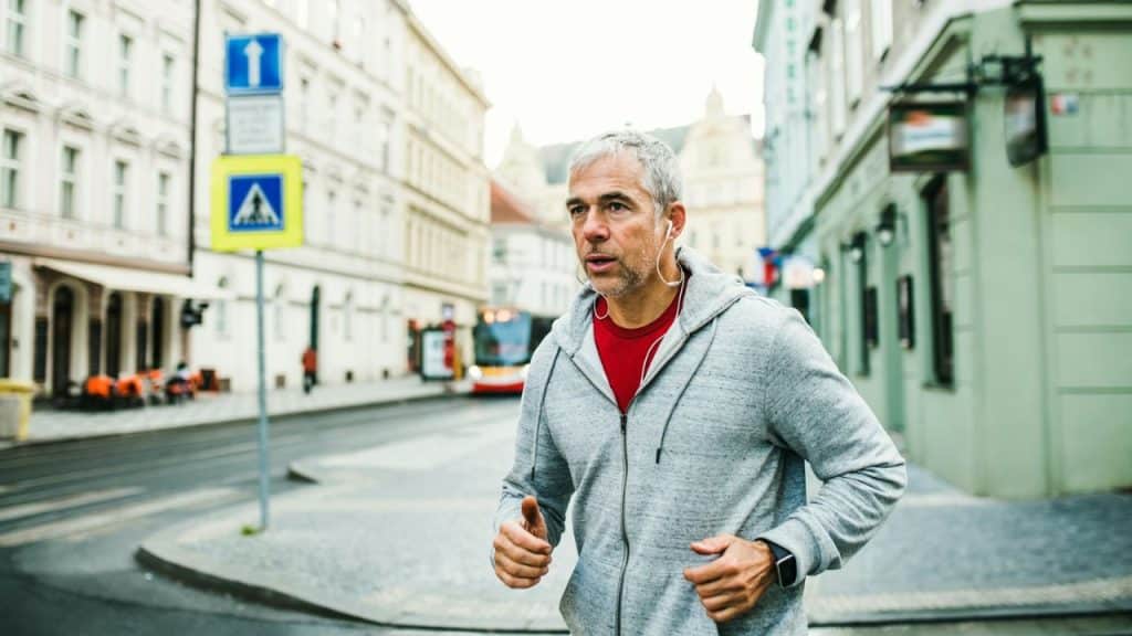 Middle-aged man jogging on a city street, wearing a gray hoodie and earphones.