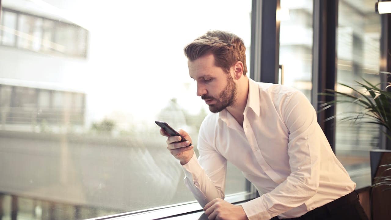 A bearded man in a white shirt is leaning by a window, looking at his smartphone.
