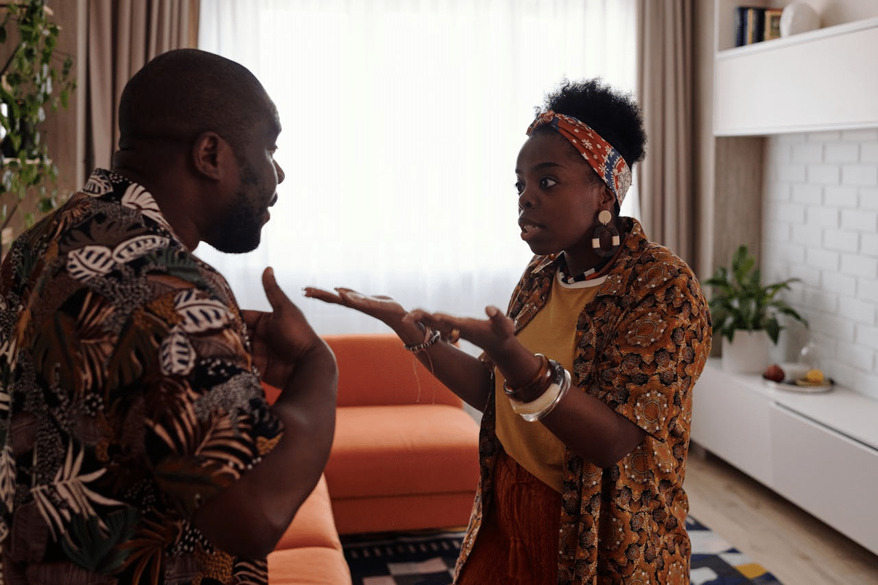 A man and woman are arguing in a living room, with the woman gesturing with open hands.