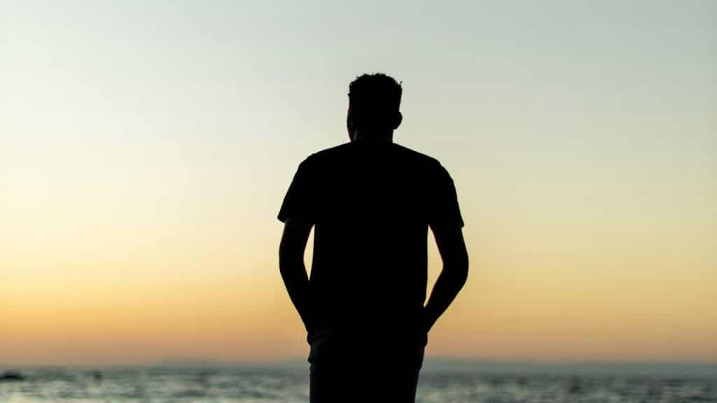 A silhouette of a man standing by the sea, looking at the sunset.