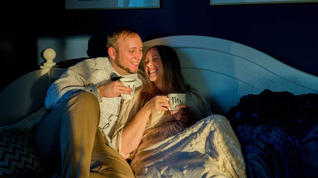 Man and woman sitting on a bed smiling and holding 'Mr.' and 'Mrs.' mugs.