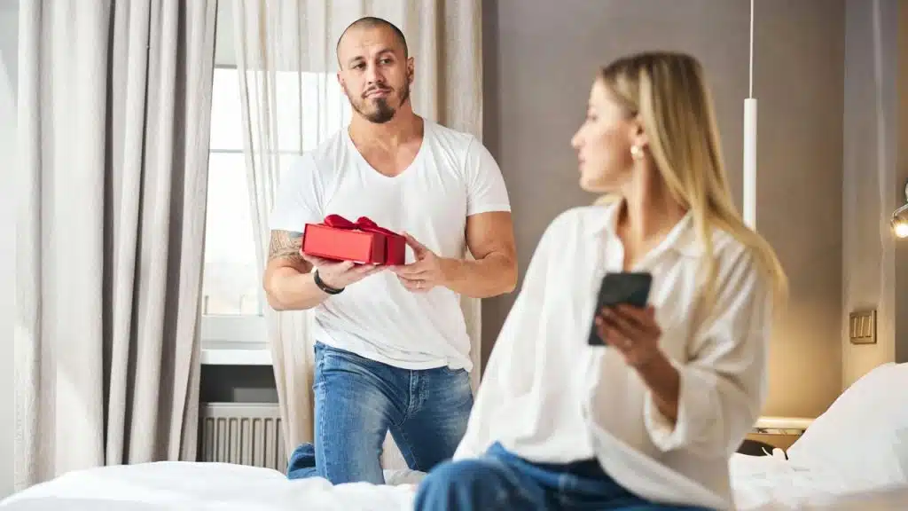 Man on one knee holding a red gift box while woman sits with a phone.