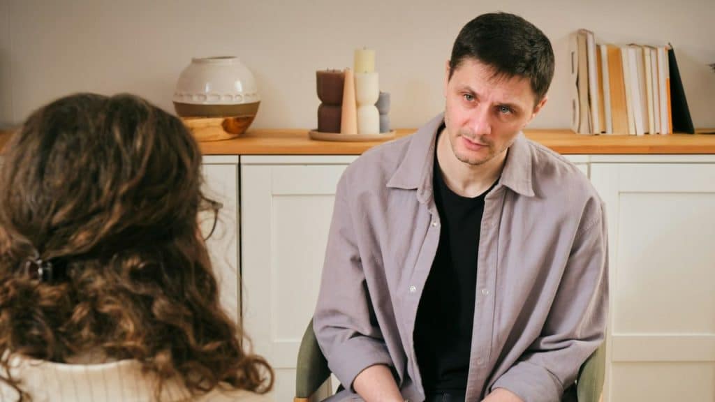 A man faces a woman with long curly hair in a bright, sparsely decorated room.