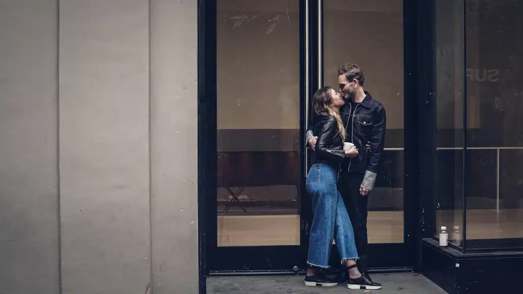 A man and a woman in dark clothing are kissing while standing outside a store window in an urban setting.