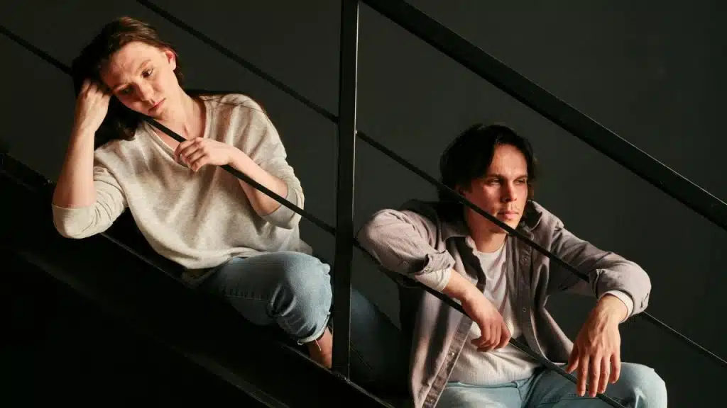 A distressed woman and a pensive man sit separately on a dark metal staircase railing.