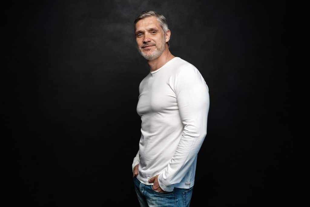 A middle-aged man in white t-shirt is posing in front of a black background.