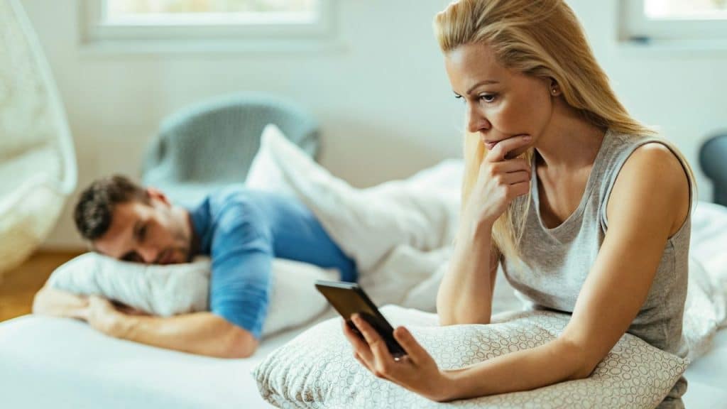 A woman checks her phone in the foreground while a man lies in bed behind her.©Curated Lifestyle/Unsplash.com