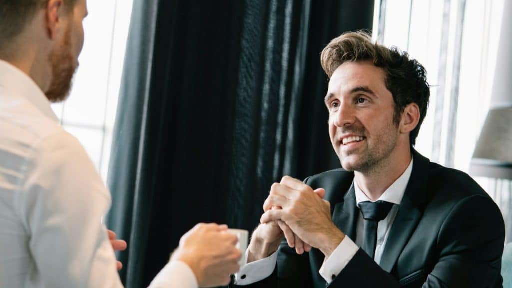 A man in a suit smiles and talks to another man across a table.