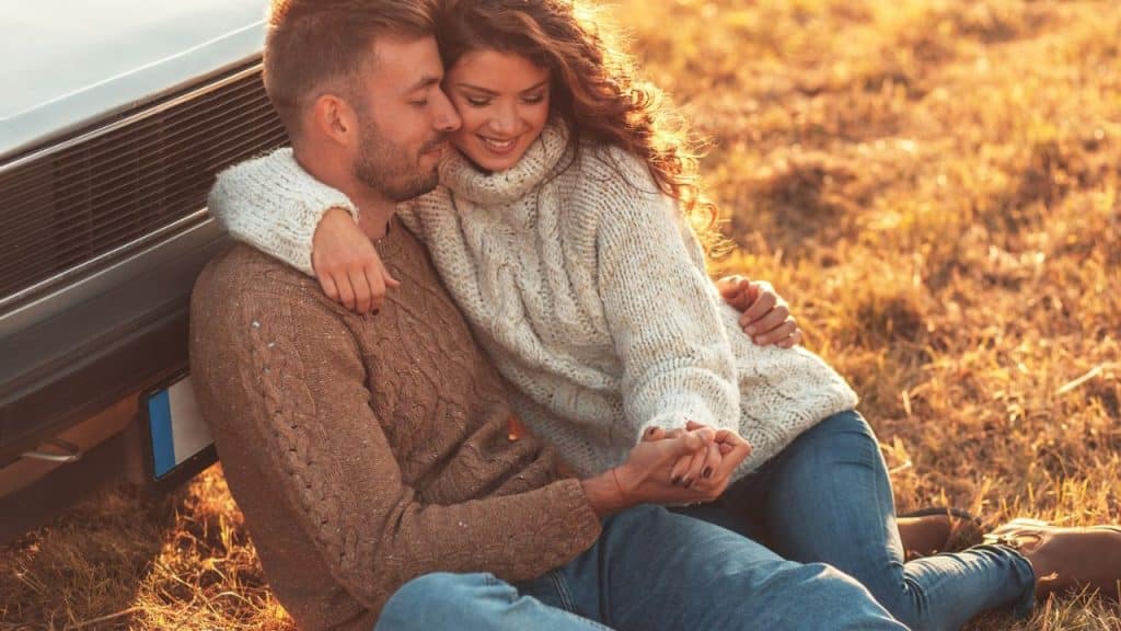 Man and woman sitting together in the sun, hugging and holding hands by a car.