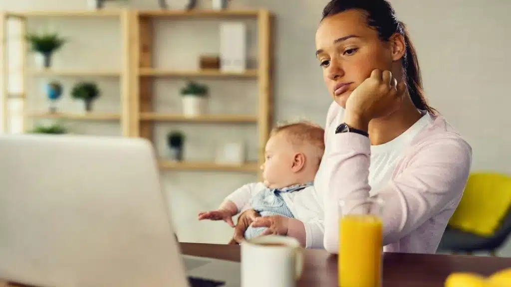 A tired woman holding a baby looks at a laptop screen in a home setting.