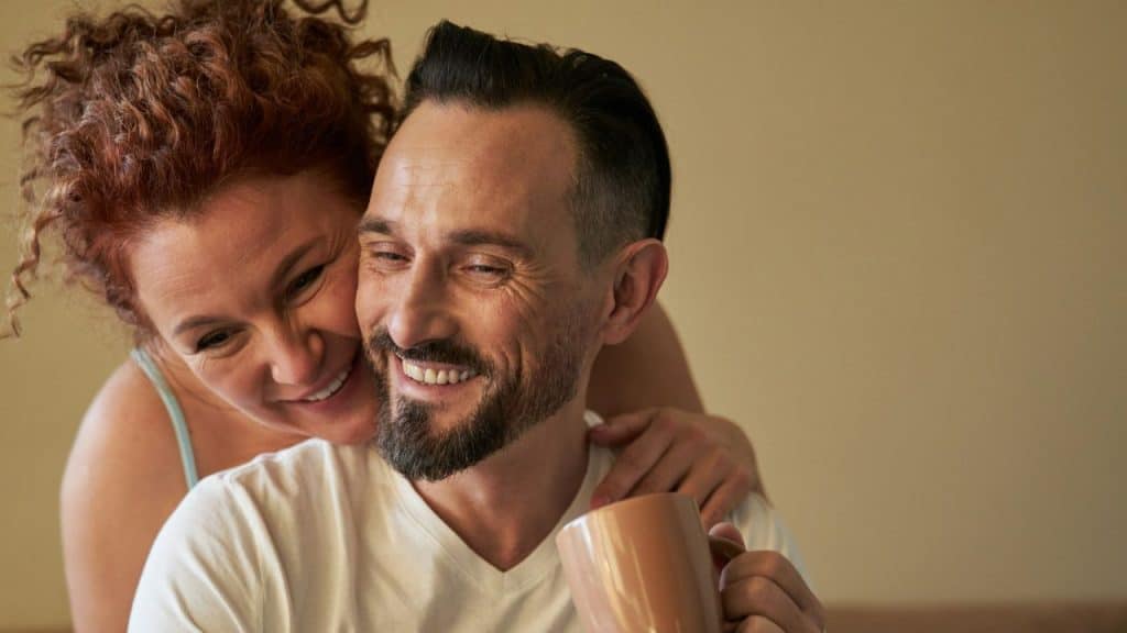 A smiling woman embraces a laughing bearded man holding a mug from behind.