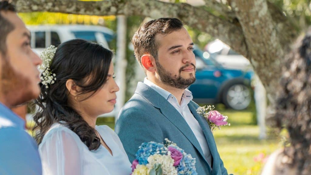 A bride and groom are sitting at an outdoor ceremony, with the groom in a blue suit and the bride in white, both looking off to the side.