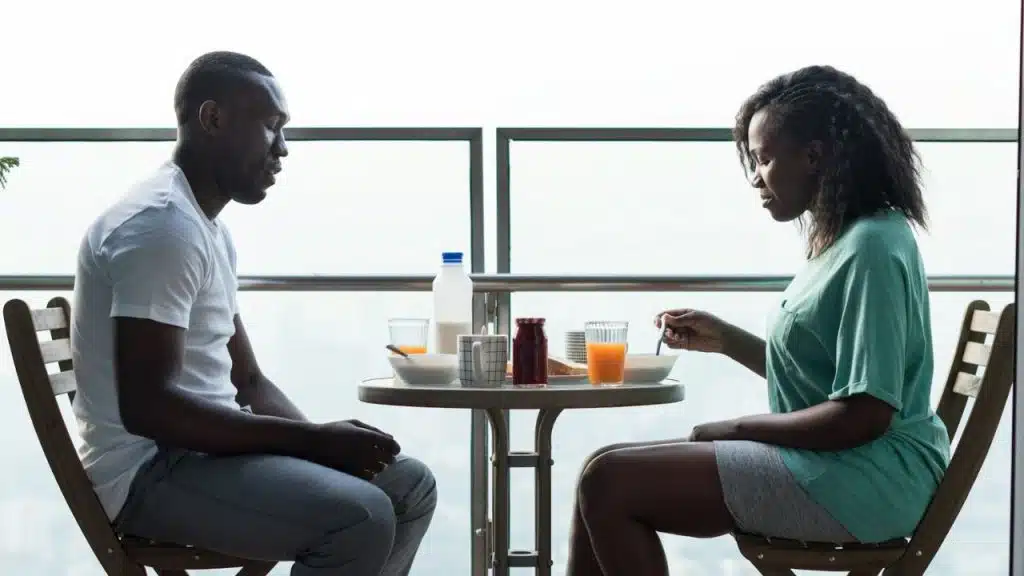 A Black man and woman are seated across a small, round table, eating breakfast on a balcony.