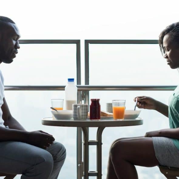 A Black man and woman are seated across a small, round table, eating breakfast on a balcony.