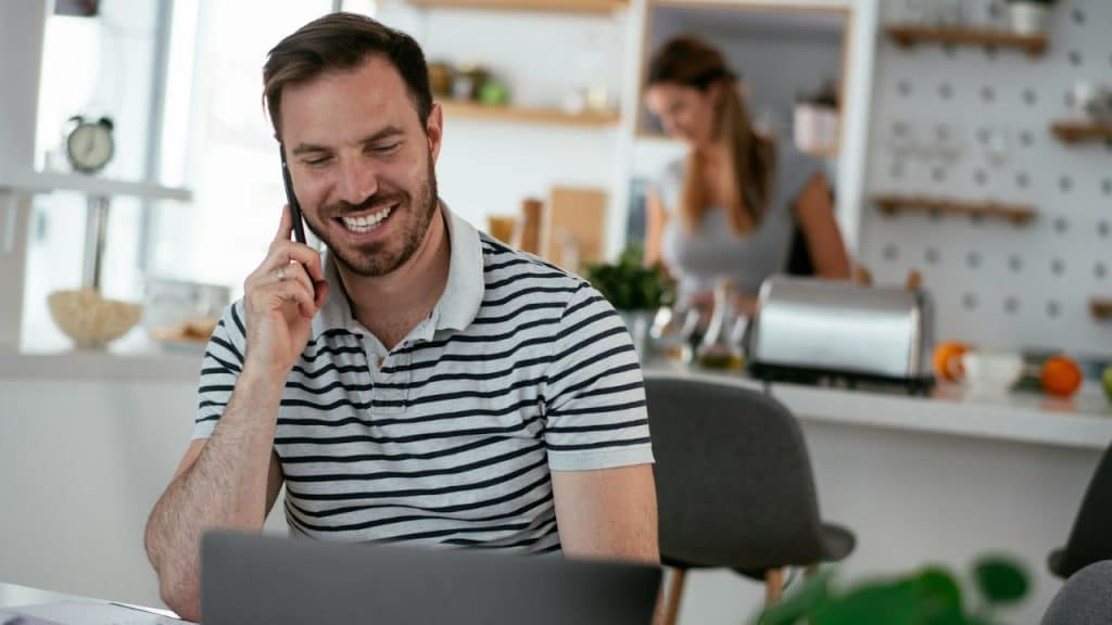 A smiling man on a phone and laptop, with a woman working in the blurry background.