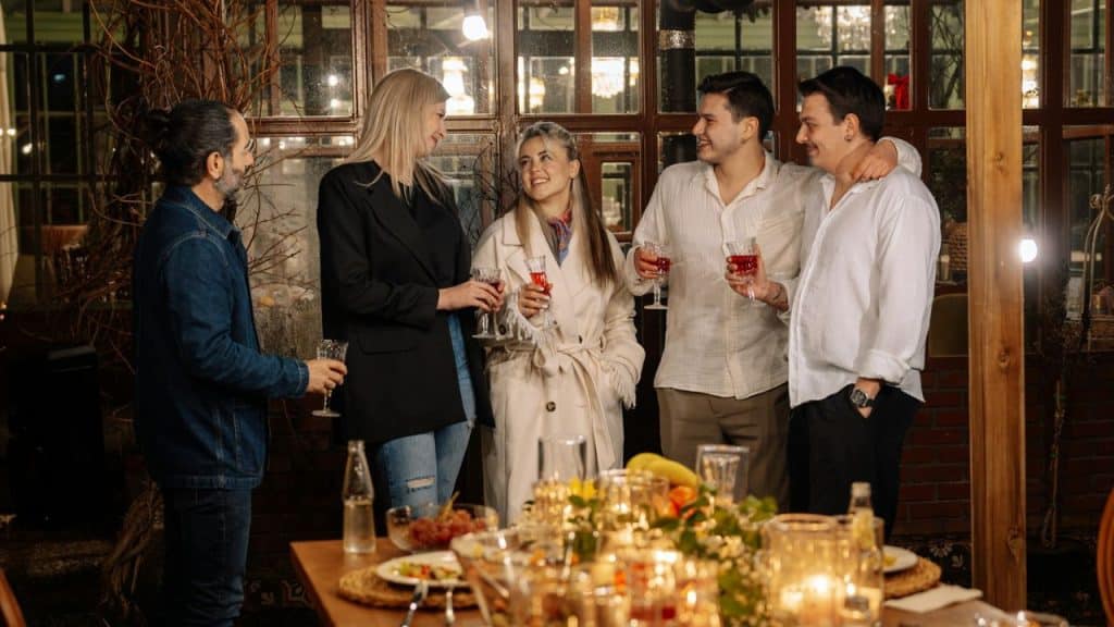 Group of five adults chatting and holding drinks at a dimly lit indoor dinner party.
