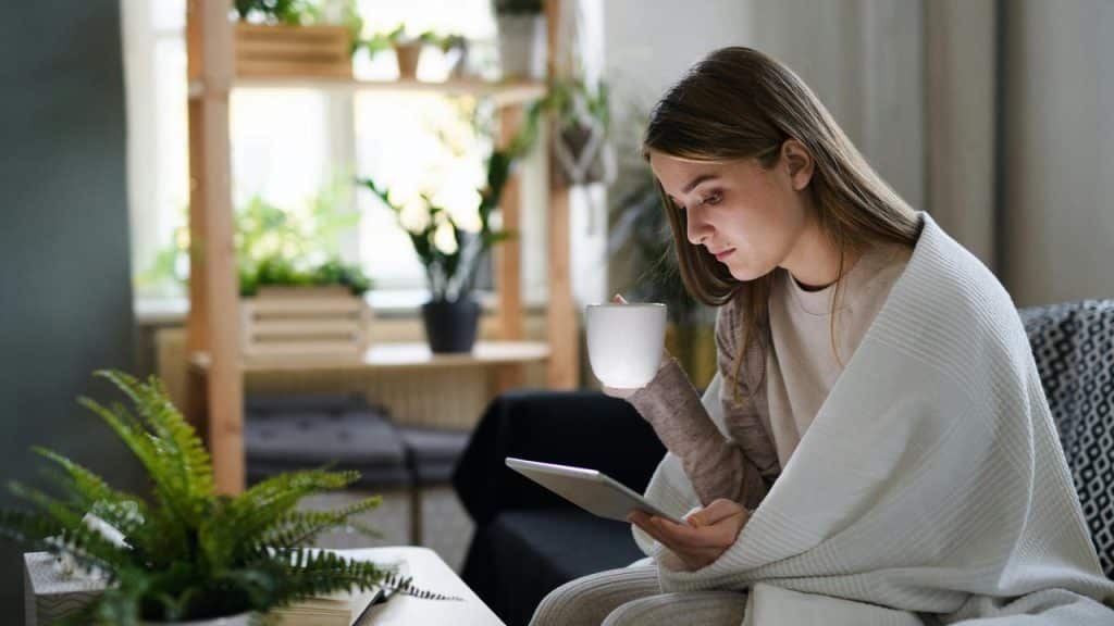 A woman wrapped in a blanket holding a mug and using a tablet.