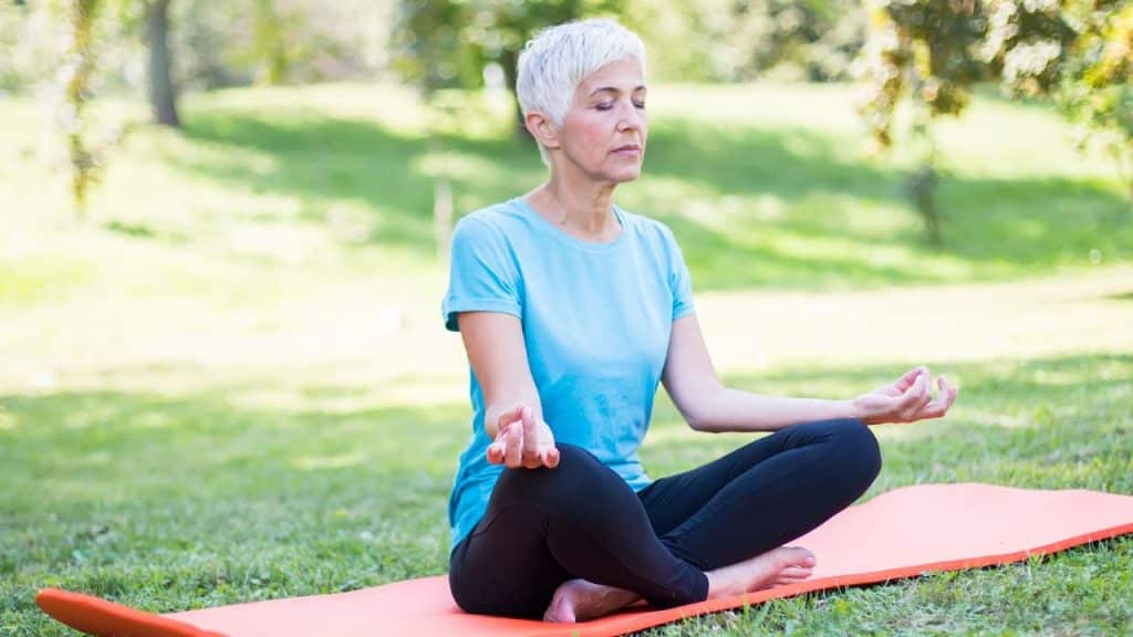 An older woman meditates in a lotus position on a yoga mat outdoors.
