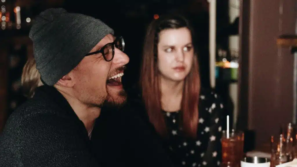 Man in beanie and glasses laughing heartily; a woman looks on in the background.
