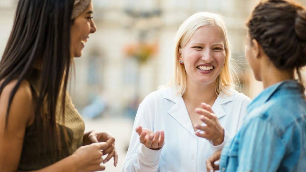 Blonde woman outdoors laughing and gesturing while talking to two other women.