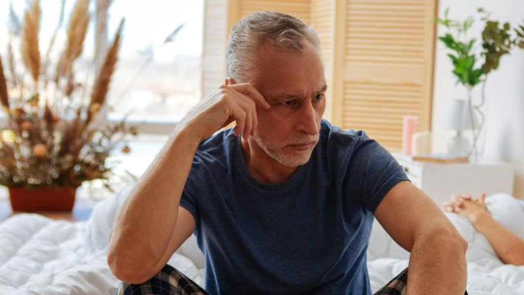 Older man sitting in bed, looking troubled, resting his head on his hand.