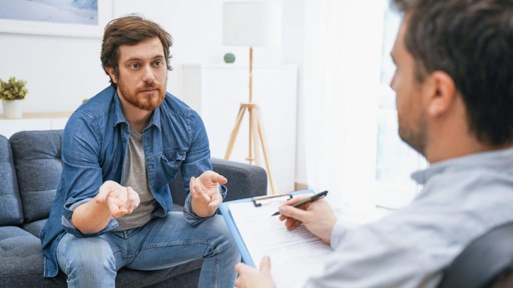 A bearded man talking to a therapist in his office.