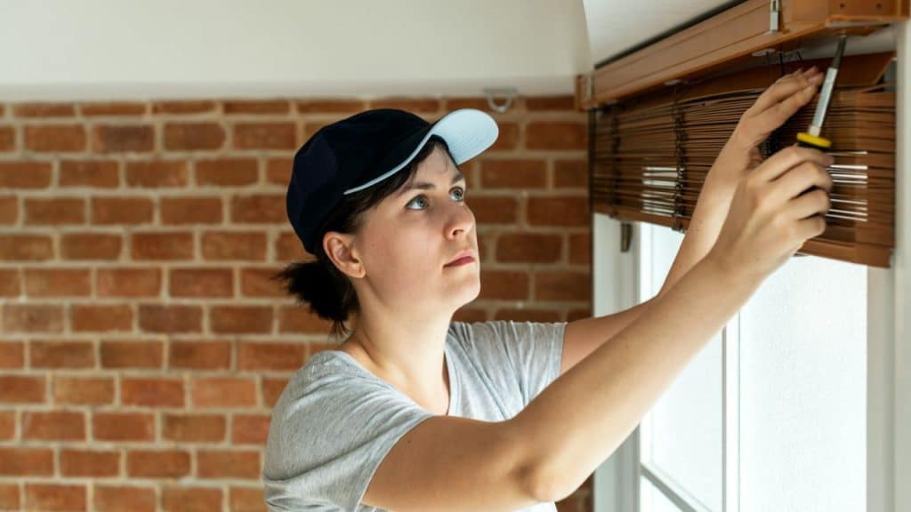 A woman with a baseball cap is using a screwdriver to fix a wooden window blind.