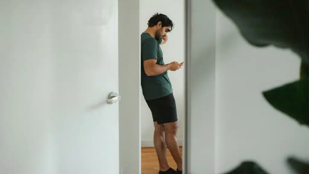 A man stands alone in a hallway looking at his phone, seen through a partially open doorway.
