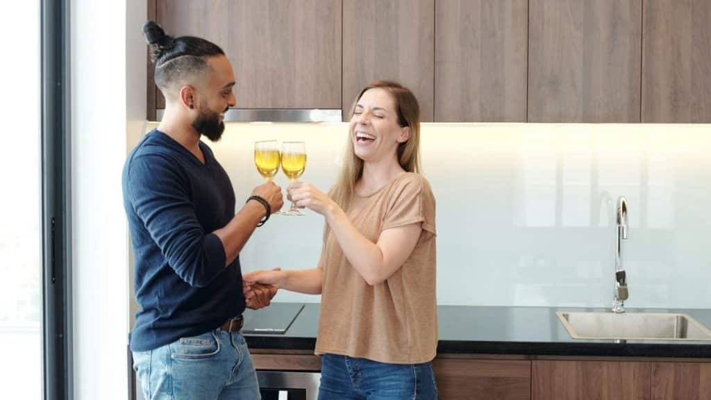 A man and a woman in a kitchen are holding up wine glasses and laughing.