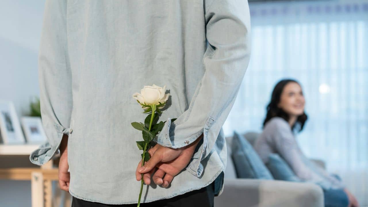 A man hides a white rose behind his back for a woman smiling nearby.