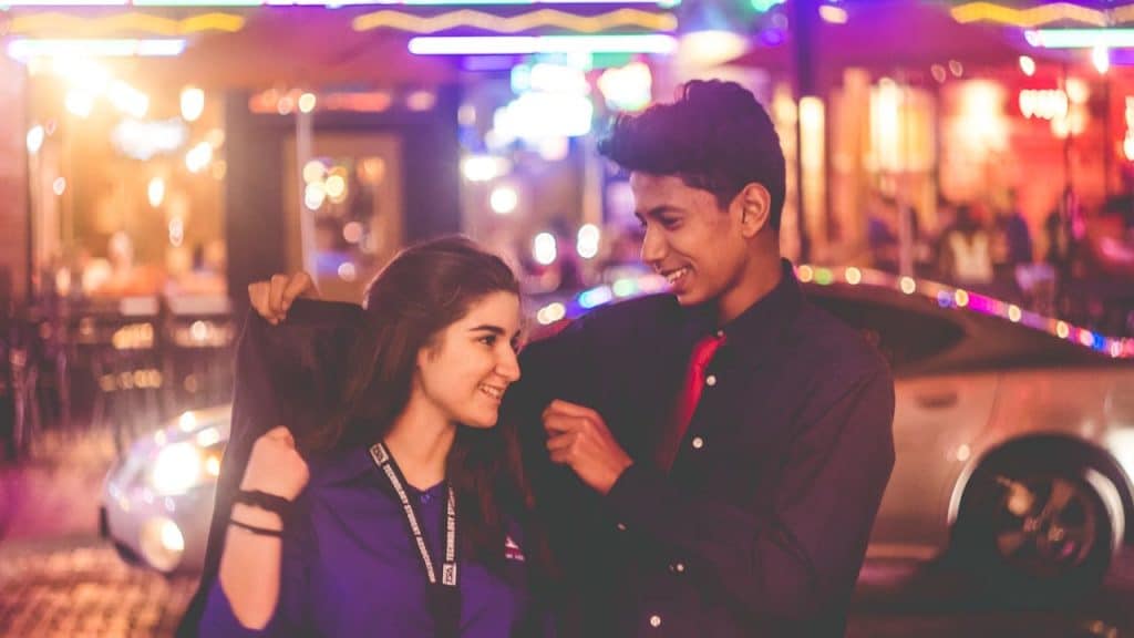 A young couple smiling at each other on a neon-lit city street at night.