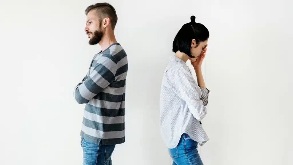 A man and woman stand back-to-back against a white wall; the woman is crying.