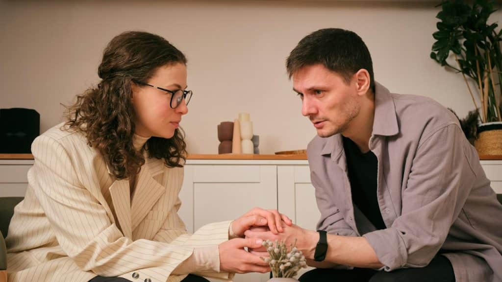 A woman wearing glasses holding the hands of and consoling an upset man.