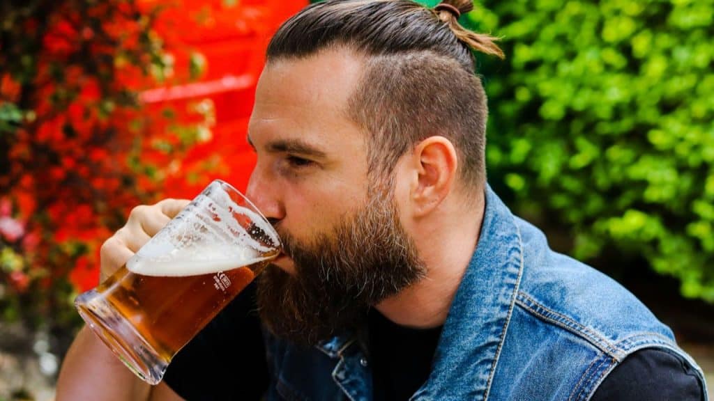 Man with beard and bun drinking beer from a mug outdoors with green and red background.