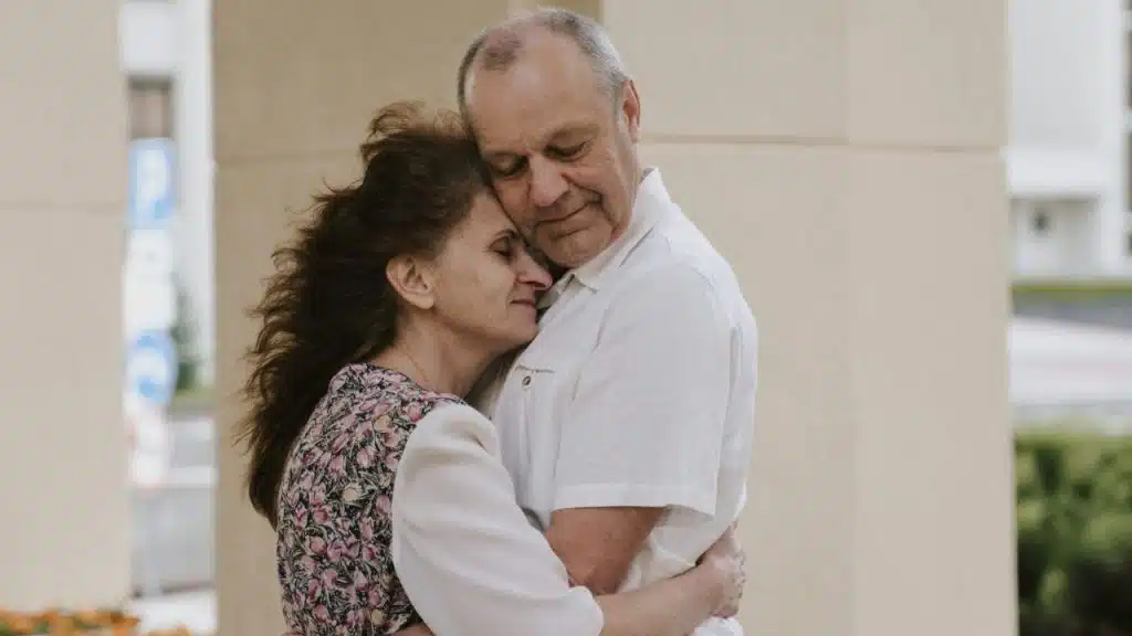 Elderly man and woman embracing outdoors with a light-colored building in the background.