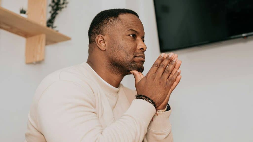 Middle-aged black man sitting indoors, hands clasped, looking thoughtfully to the side.