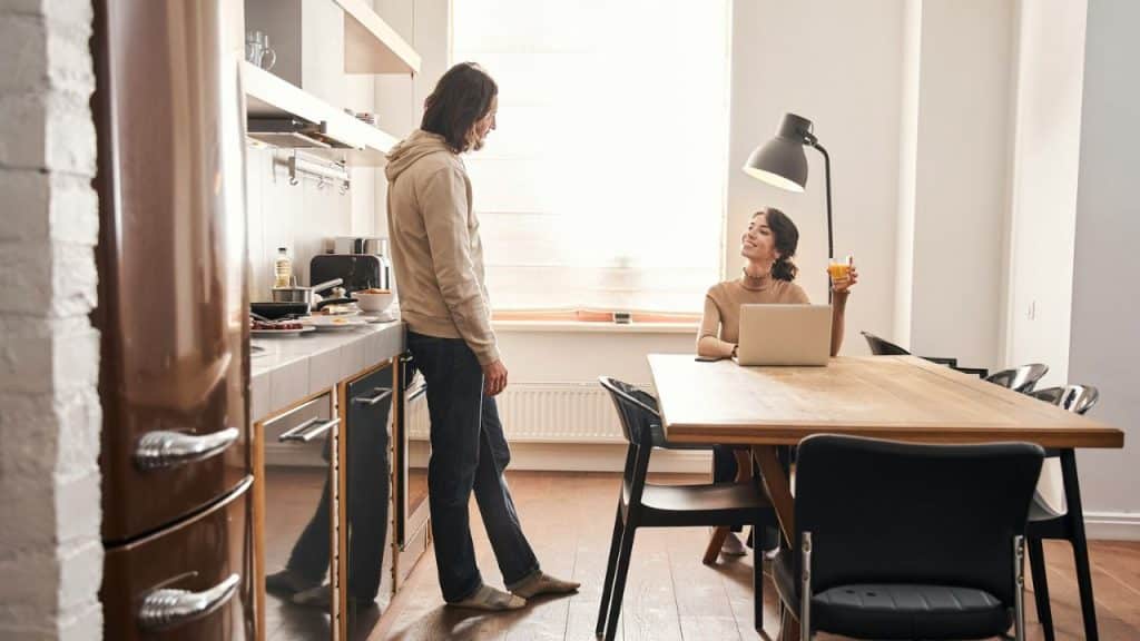 Couple in a kitchen, one standing, one sitting at a table with a laptop.