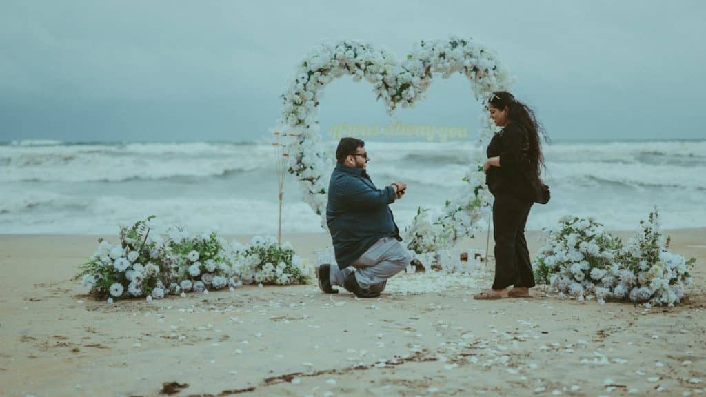 A man proposes to a woman on a beach under a heart arch.