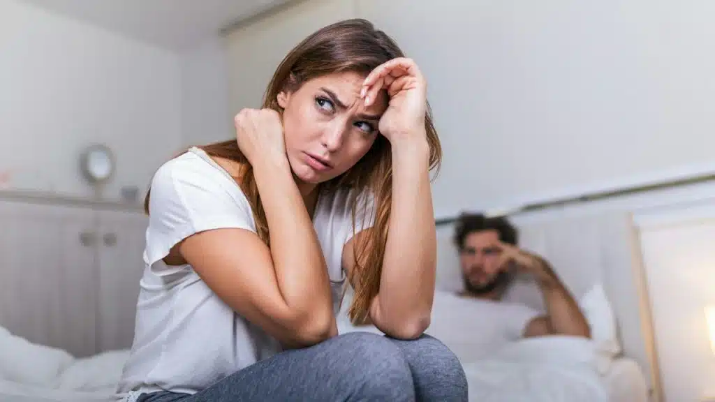 A focused woman sits on a bed with her head in her hands, while a man is blurry behind her.