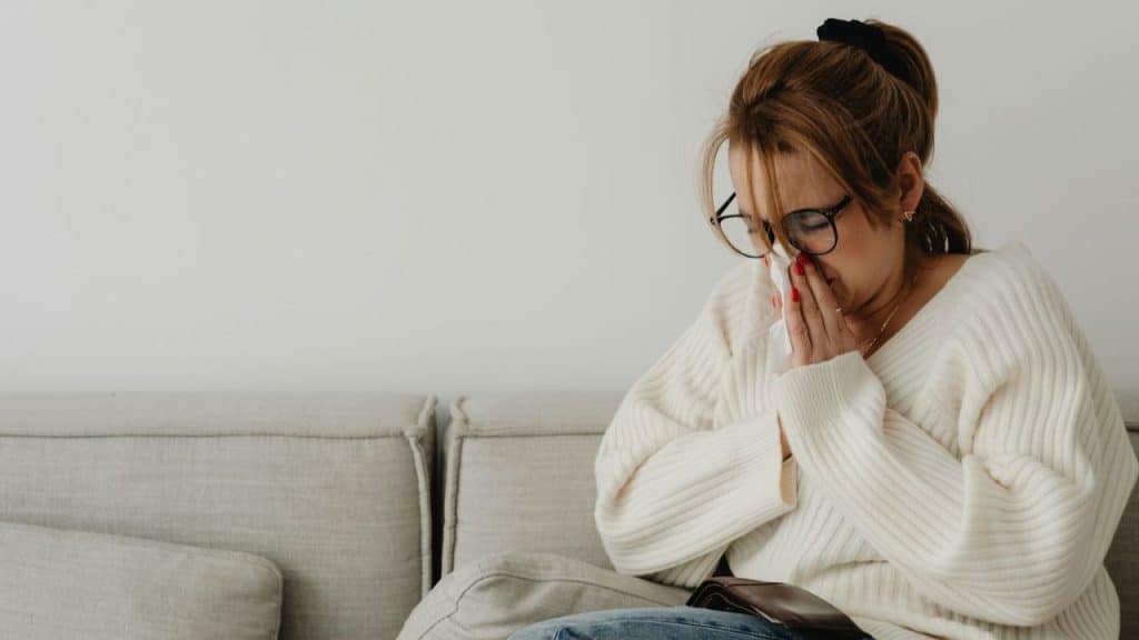 Woman in a white sweater and glasses holds a tissue to her face while sitting.
