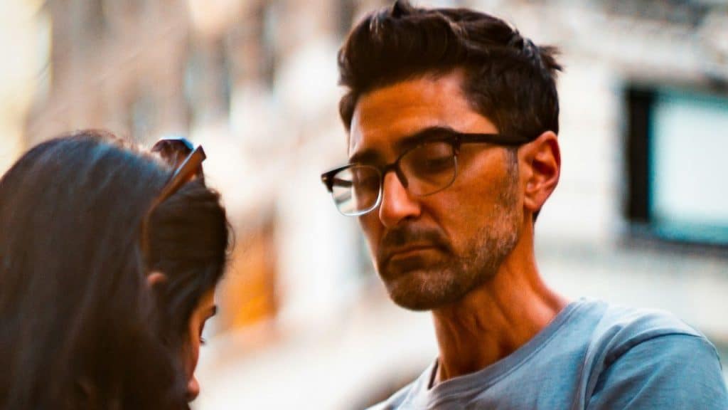 Man in glasses and gray shirt looking intently at another person's head.