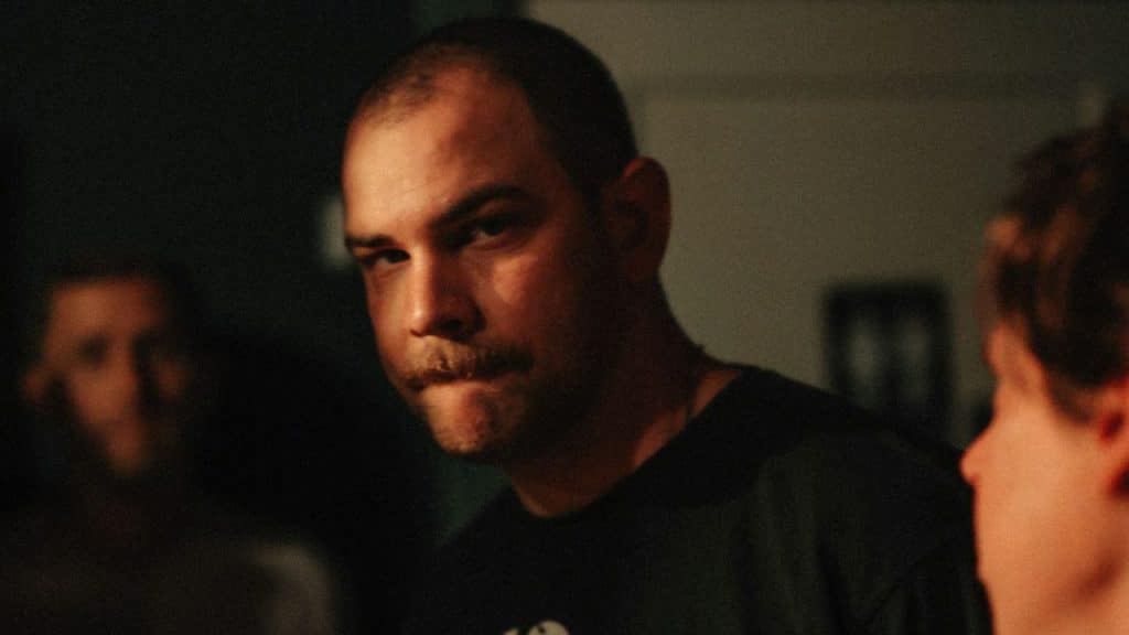 Bald man with a mustache looks intensely into the distance in a dimly lit room.