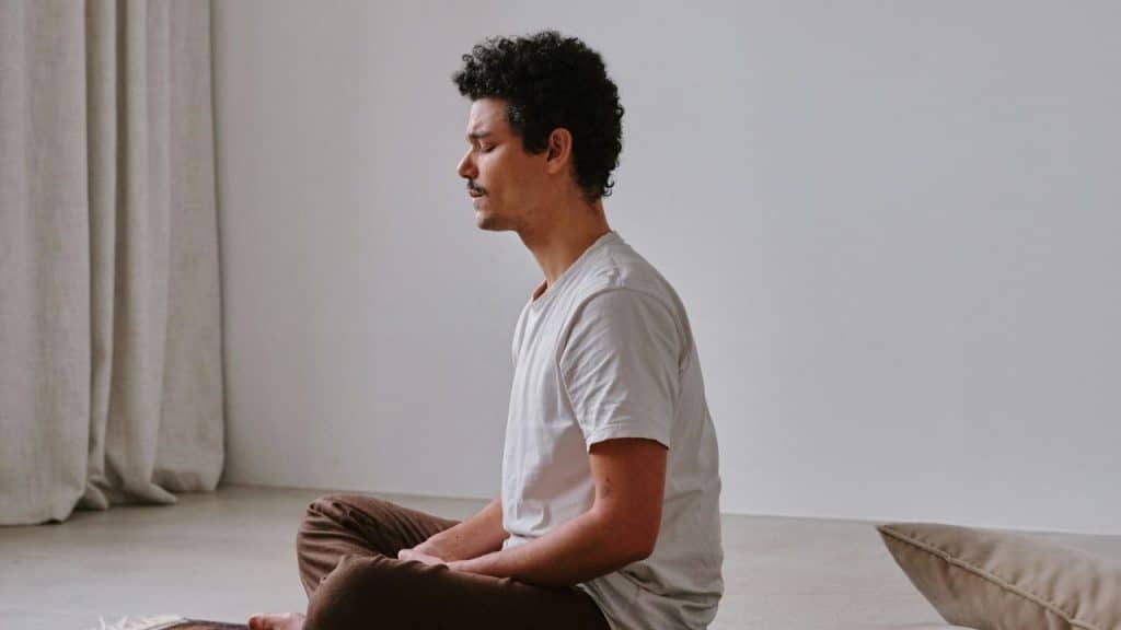 Man with mustache and curly hair sitting cross-legged, eyes closed, meditating indoors.