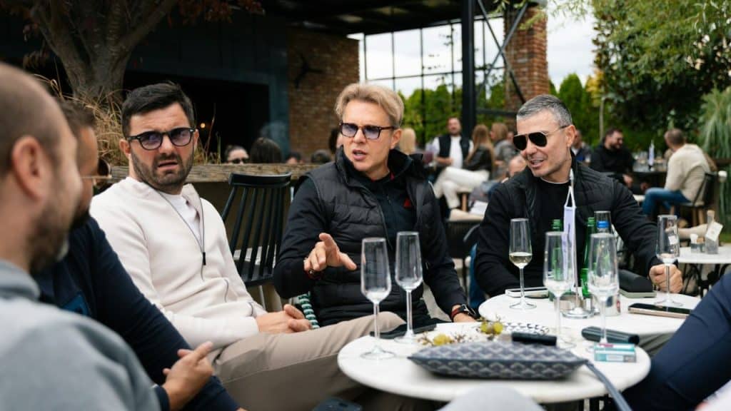 Three men sitting around a table with drinks outdoors, one is speaking.